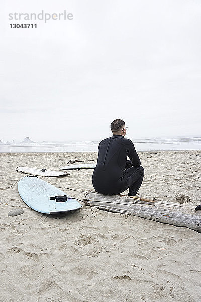 Rückansicht eines Mannes  der auf einem Baumstamm am Strand an Surfbrettern gegen den Himmel sitzt