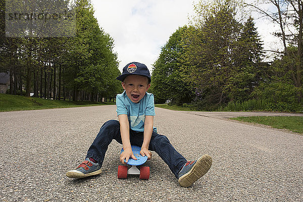 Porträt eines verspielten Jungen  der auf einem Skateboard am Fussweg sitzt