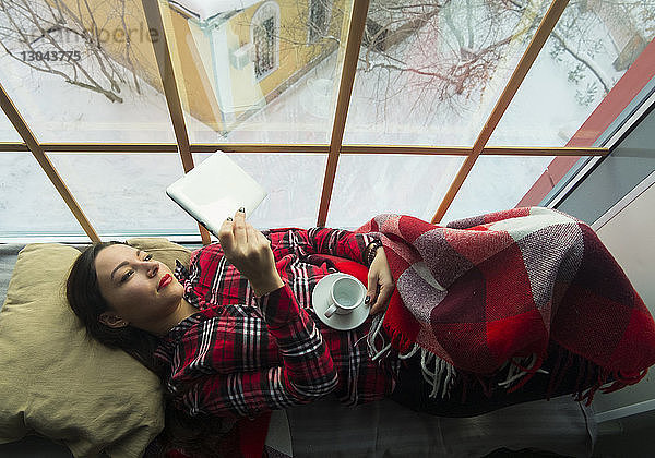 Draufsicht einer Frau  die einen Tablet-Computer benutzt  während sie sich auf einem Fensterplatz in einer Nische entspannt