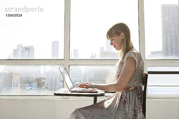 Lächelnde Geschäftsfrau mit Laptop auf Tisch gegen Fenster