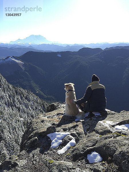 Rückansicht eines Mannes mit Hund  der auf einem Felsen gegen einen Berg sitzt