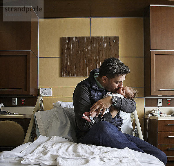 Vater küsst neugeborenen Sohn  während er ihn im Krankenhausbett trägt