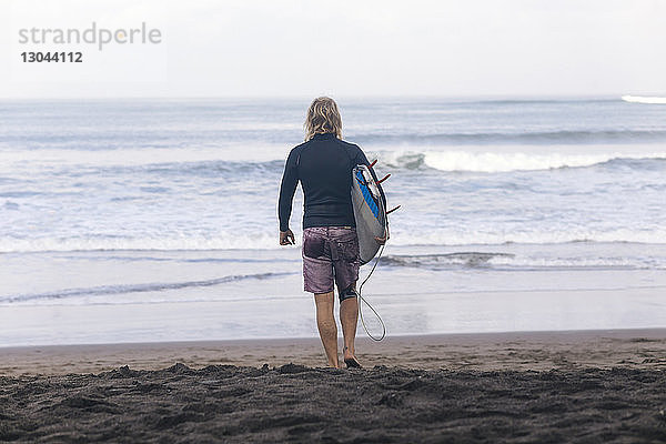 Rückansicht eines Mannes  der ein Surfbrett trägt  während er am Strand in Richtung Meer läuft