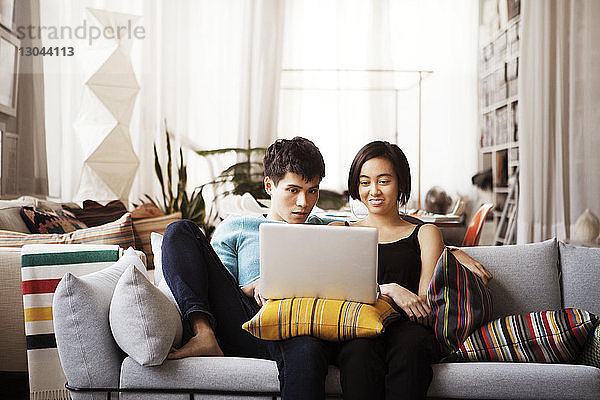 Junges Paar benutzt Laptop  während es zu Hause auf dem Sofa sitzt