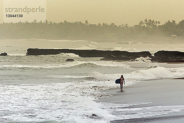Frau mit Surfbrett geht am Strand gegen den Himmel