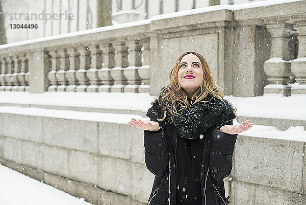 Lächelnde Frau geniesst Schnee  während sie gegen Gebäude in der Stadt steht
