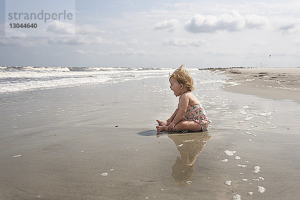 Seitenansicht eines kleinen Mädchens  das schreiend am Strand gegen den Himmel sitzt