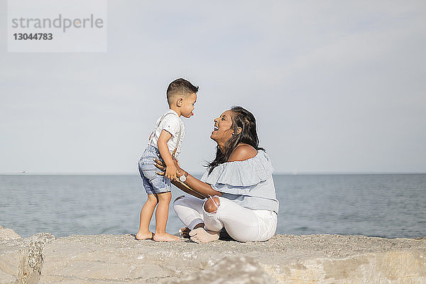 Seitenansicht eines glücklichen Sohnes und einer glücklichen Mutter  die spielen  während sie auf einer Stützmauer gegen das Meer sitzen