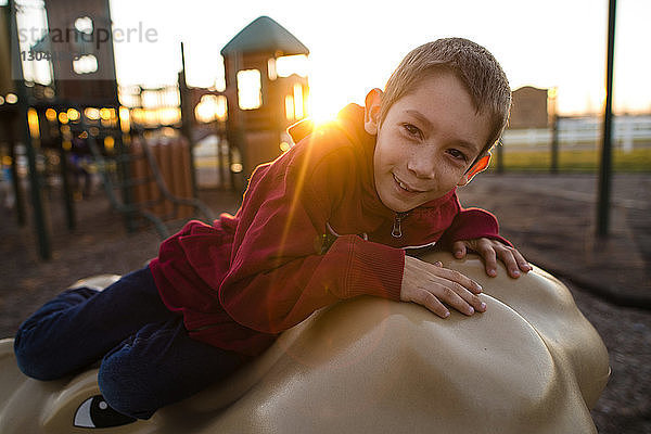 Porträt eines glücklichen Jungen  der bei Sonnenuntergang auf einer Tierstatue auf dem Spielplatz spielt