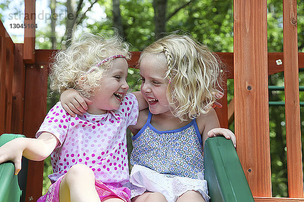 Fröhliche Schwestern schauen sich Gesicht zu Gesicht  während sie auf einer Rutsche auf dem Spielplatz sitzen