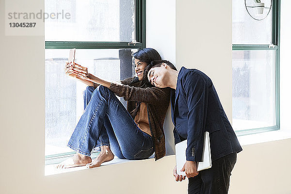 Fröhliche Geschäftsfrau nimmt Selfie mit Kollegen  die im Kreativbüro auf der Schulter schlafen