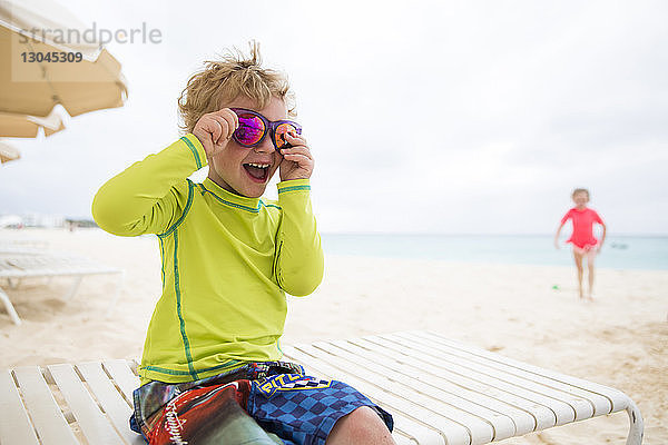Verspielter Junge mit glänzender Sonnenbrille  der am Strand auf einem Liegestuhl sitzt