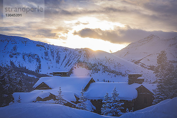 Szenische Ansicht von Häusern an schneebedeckten Bergen vor bewölktem Himmel bei Sonnenaufgang