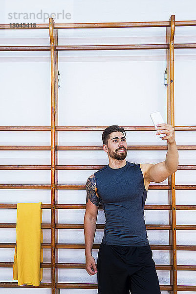 Lächelnder  muskulöser Athlet  der sich mit seinem Smartphone gegen Sprossenwände im Fitnessstudio drückt