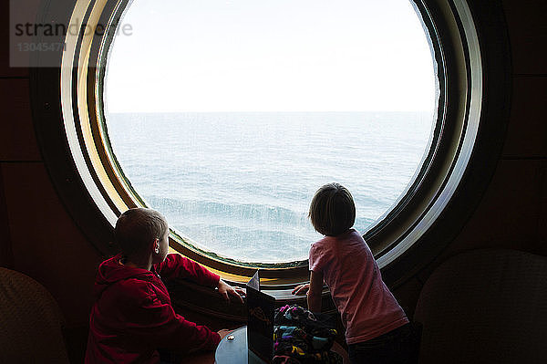 Geschwister schauen aufs Meer  während sie am Bullauge eines Kreuzfahrtschiffes stehen