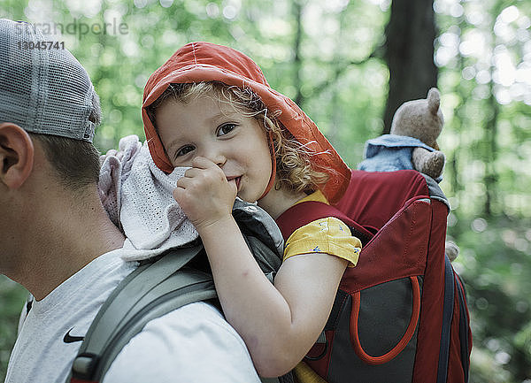 Porträt einer süßen glücklichen Tochter  die am Daumen lutscht  während sie vom Vater im Wald getragen wird