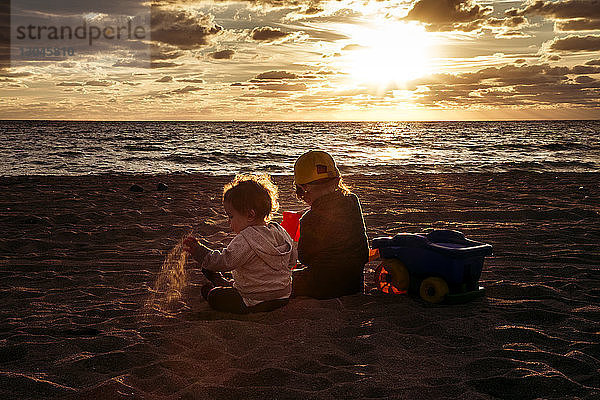 Rückansicht von Geschwistern  die bei Sonnenuntergang auf Sand am Strand gegen den Himmel sitzen