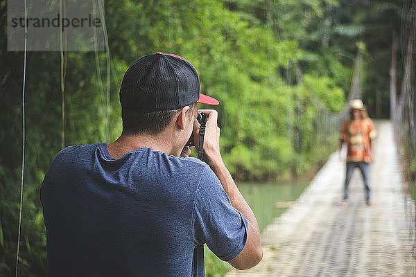 Rückansicht eines Mannes  der einen Freund fotografiert  während er auf einer Seilbrücke steht