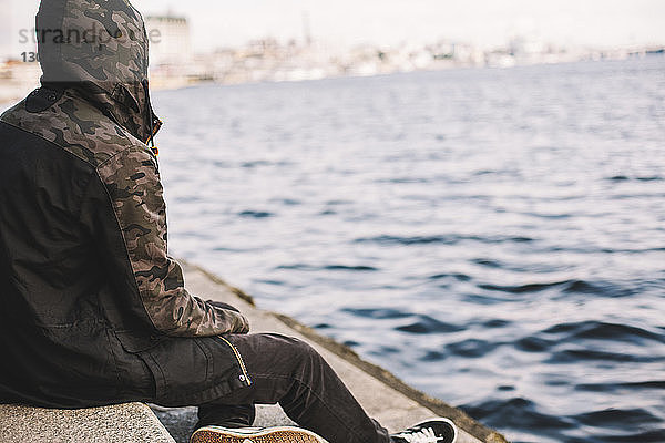 Junger Mann trägt Kapuzenjacke  während er auf Stufen am Flussufer sitzt