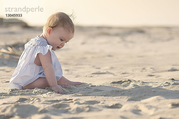 Seitenansicht eines kleinen Mädchens  das bei Sonnenuntergang auf Sand am Strand gegen den Himmel sitzt