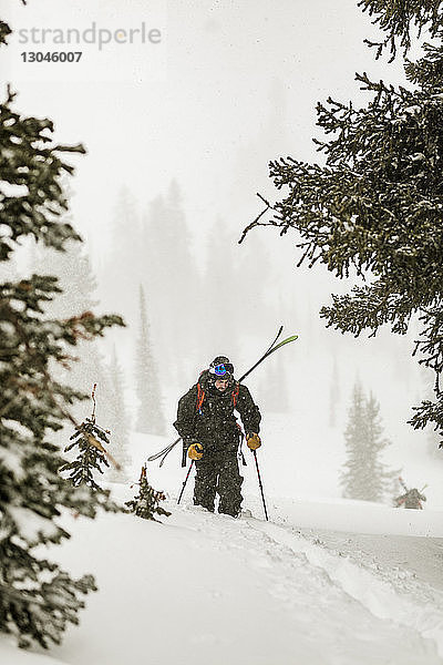 Mann mit Skiern geht bei Schneefall auf schneebedecktem Feld im Wald