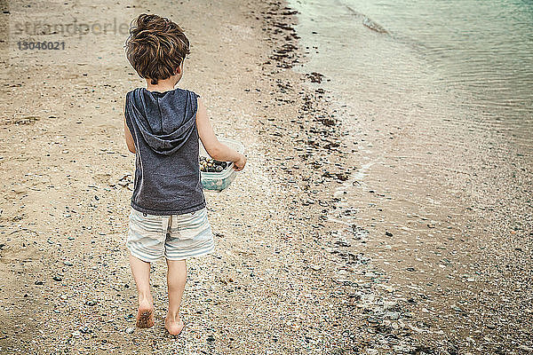 Rückansicht eines Jungen  der beim Strandspaziergang eine Schale mit Kieselsteinen trägt