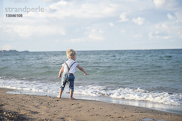 Rückansicht eines kleinen Jungen  der am Strand vor wolkenverhangenem Himmel stehend einen Stein abschöpft