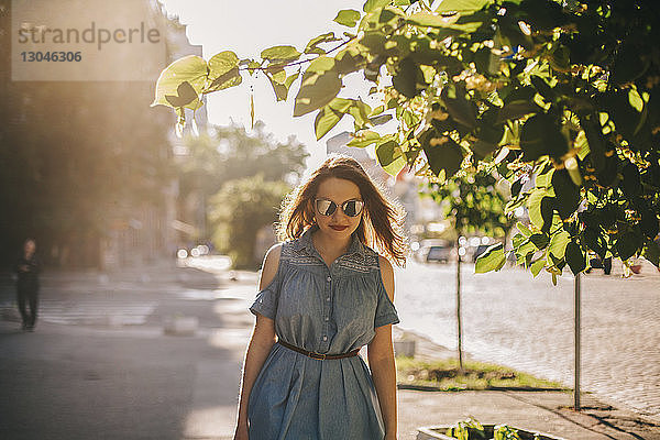 Junge Frau mit Sonnenbrille geht bei Sonnenschein in der Stadt auf dem Bürgersteig