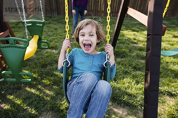 Porträt eines glücklichen Mädchens  das auf einem Spielplatz mit der Mutter im Hintergrund schaukelt