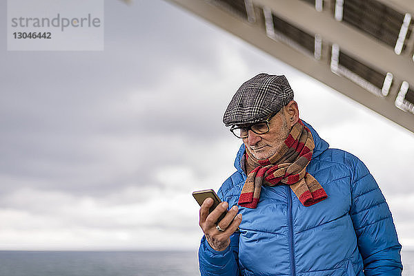 Älterer Mann benutzt Smartphone  während er am Meer vor bewölktem Himmel steht