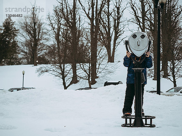 Junge blickt durch ein Münzfernglas auf verschneites Feld gegen kahle Bäume