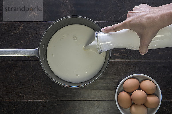 Ausgeschnittenes Bild von Händen  die Milch in der Pfanne nach Eiern auf den Tisch gießen