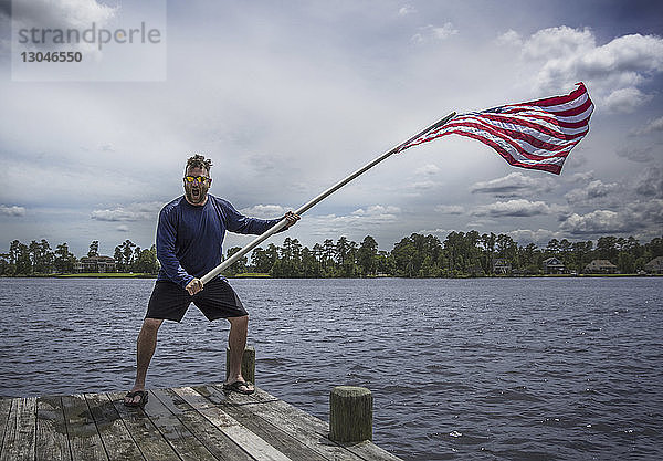 Fröhlicher Mann schwenkt die amerikanische Flagge  während er auf dem Pier über dem See vor bewölktem Himmel im Banff-Nationalpark steht