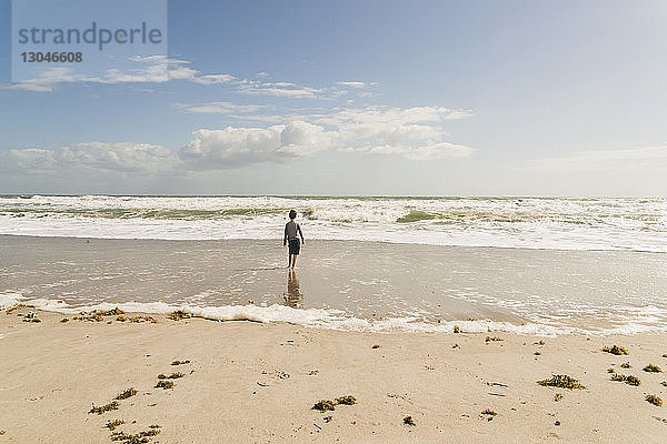 Rückansicht eines Jungen  der bei Sonnenschein am Strand gegen den Himmel steht