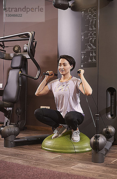 Frau  die mit Widerstandsbändern trainiert  während sie in der Hocke auf dem Balance-Trainingsball im Fitnessstudio kauert