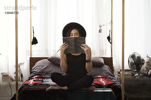Glückliche Frau versteckt Gesicht mit Buch  während sie zu Hause auf dem Bett sitzt