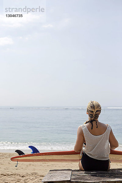 Rückansicht einer am Strand sitzenden Frau mit Surfbrett gegen den Himmel