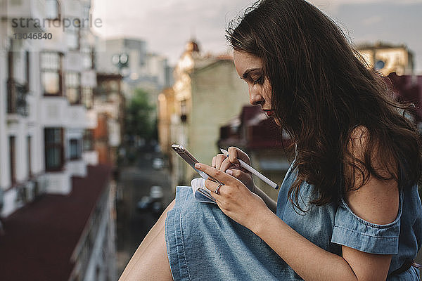 Seitenansicht einer jungen Frau  die ein Smartphone benutzt  während sie an Gebäuden in der Stadt sitzt