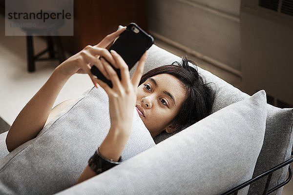 Hochwinkelansicht einer jungen Frau  die ein Smartphone benutzt  während sie zu Hause auf dem Sofa liegt