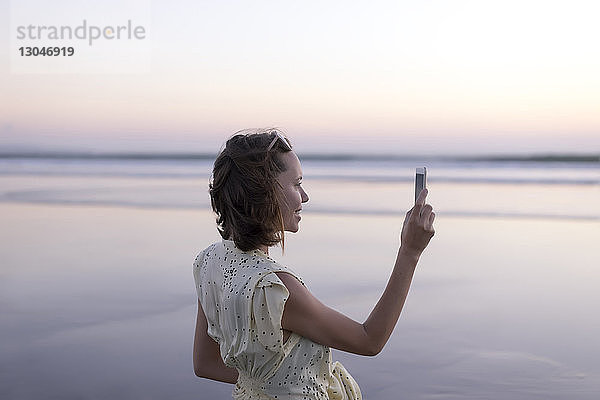 Seitenansicht einer glücklichen Frau  die mit einem Smartphone fotografiert  während sie am Strand gegen den Himmel steht