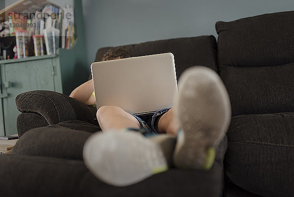 Ein Junge in voller Länge benutzt einen Laptop-Computer  während er sich zu Hause auf dem Sofa entspannt