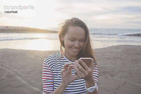 Lächelnde Frau benutzt Mobiltelefon am Strand bei Sonnenuntergang