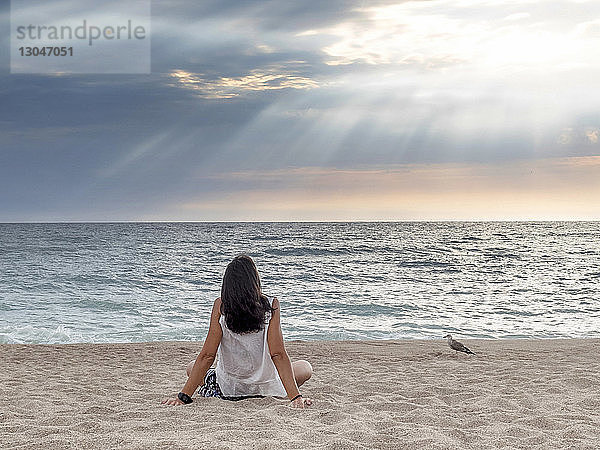 Rückansicht einer Frau  die auf das Meer schaut  während sie sich im Sonnenuntergang auf Sand vor bewölktem Himmel entspannt
