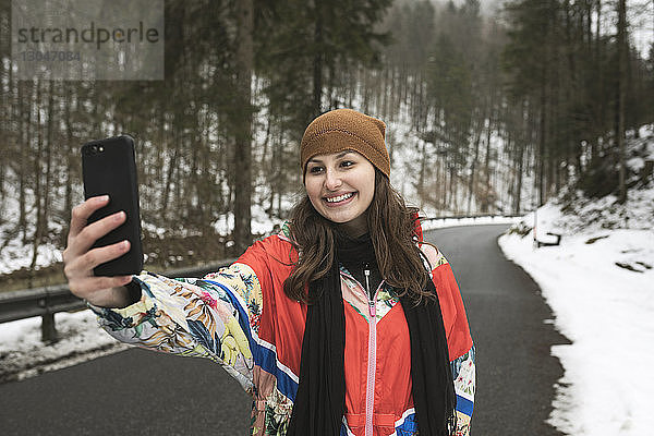 Lächelnde junge Frau  die sich mit einem Smartphone selbstständig macht  während sie im Winter auf einer Landstraße im Wald steht