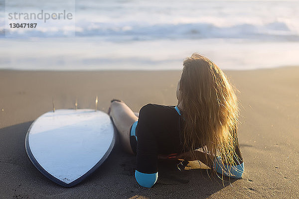 Rückansicht einer Frau  die sich bei Sonnenuntergang am Strand am Surfbrett entspannt