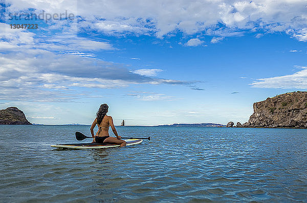 Frau sitzt auf einem Paddelbrett im Meer vor blauem Himmel