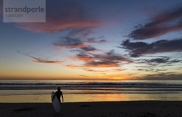 Rückansicht eines Mannes mit Surfbrett  der bei Sonnenuntergang am Strand gegen den Himmel läuft