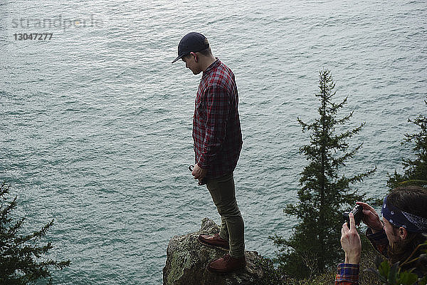 Hochwinkelaufnahme eines Mannes  der einen Freund fotografiert  der auf einem Felsen am Meer steht