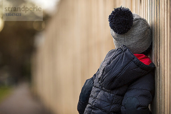 Seitenansicht eines Jungen in warmer Kleidung  der sich im Hinterhof an einen Holzzaun lehnt