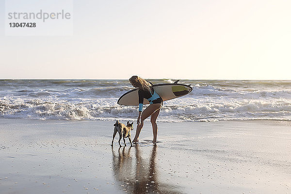 Frau in voller Länge trägt Surfbrett  während sie mit Hund am Strand steht